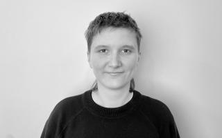 Person mit kurzem Haar trägt einen dunklen Pullover vor weißem Hintergrund.