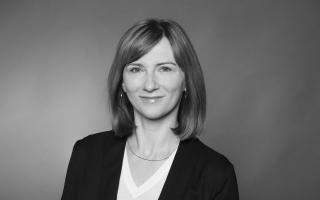 Frau mit schulterlangem, glattem Haar trägt ein schwarzes Sakko und ein weißes V-Ausschnitt-Oberteil vor einfarbigem Hintergrund.