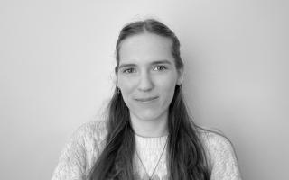 Portrait von Person mit langem, glattem Haar, die einen Pullover und eine Halskette trägt, vor einfarbigem Hintergrund, schwarz-weiß.