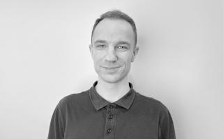 Person mit kurzen Haaren trägt ein dunkelgraues Poloshirt mit Knopfleiste vor weißer Wand.