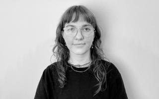 Person mit schulterlangem, welligem Haar trägt ein schwarzes T-Shirt und zwei Ketten um den Hals vor weißem Hintergrund.