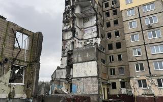 Zerstörtes mehrstöckiges Wohngebäude mit freigelegten Innenwänden und Trümmern, daneben teilweise eingestürztes Gebäude, bewölkter Himmel.